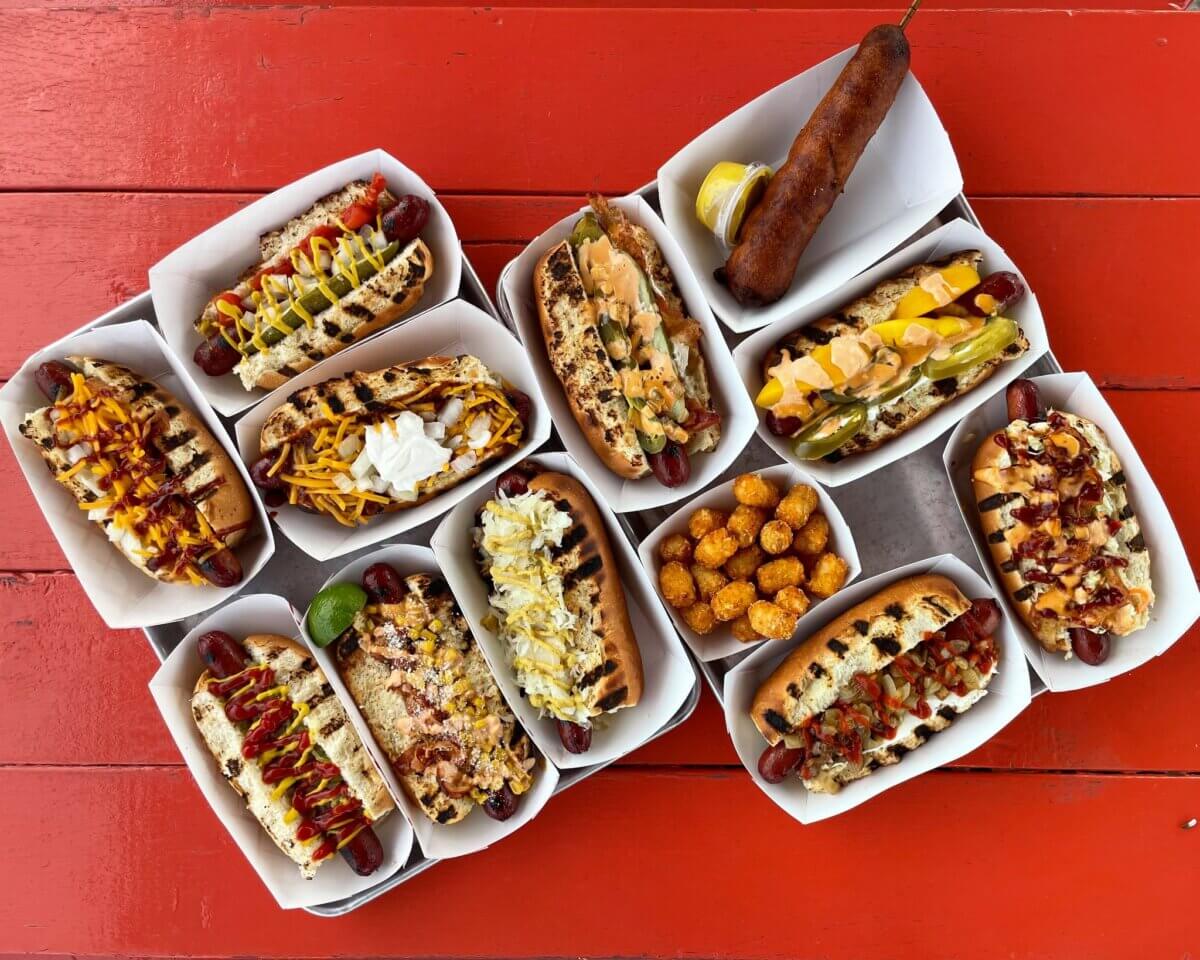 A variety of loaded hot dogs in trays, including a spicy Nashville hot dog, a corn dog, and a serving of tater tots arranged on a red wooden table.