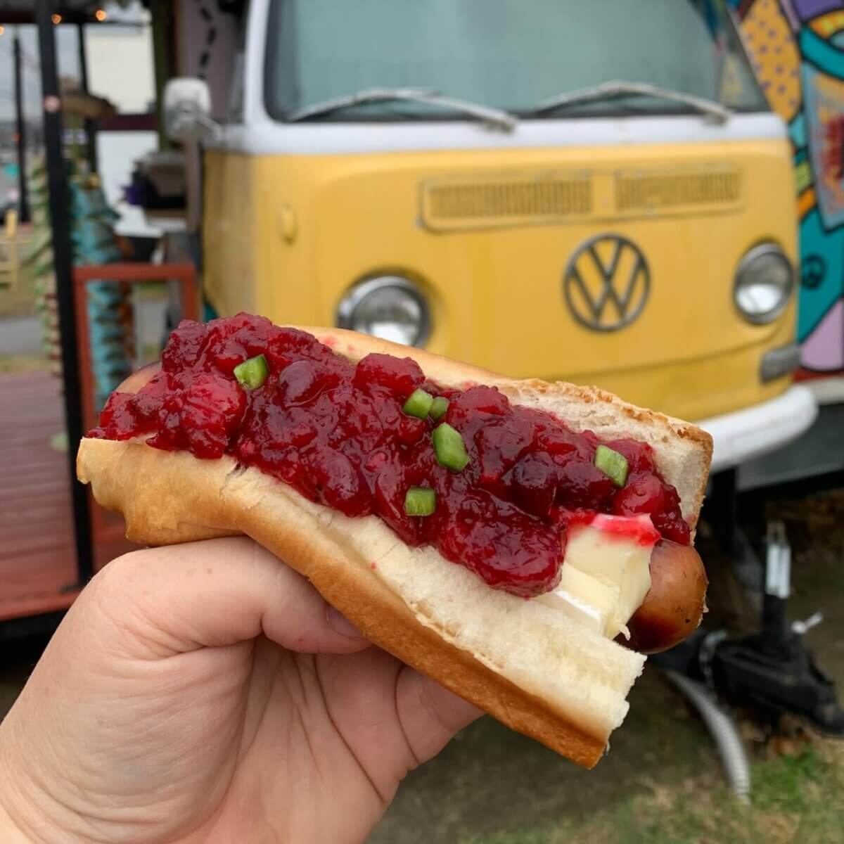 A hand holds a Nashville hot dog topped with brie cheese, cranberry sauce, and green onions in front of a yellow VW van food truck, ready to take orders.