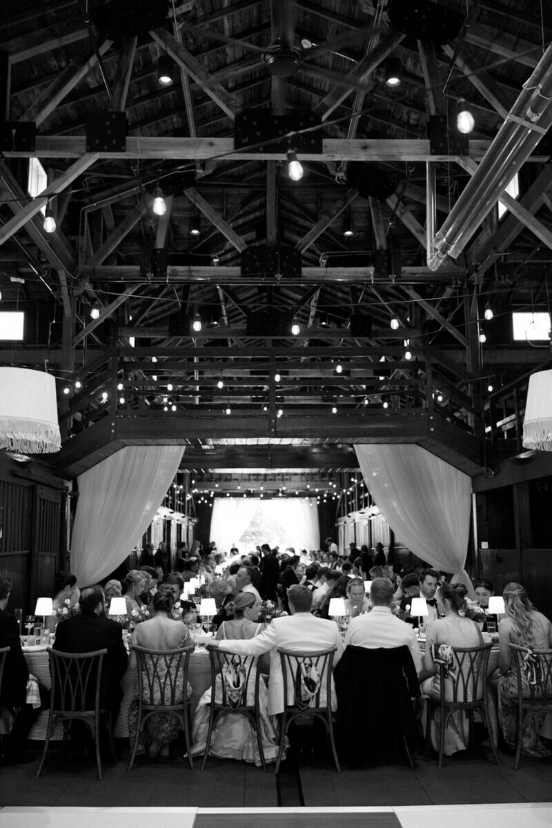 This Dreamy Equestrian Wedding Made History at Franklin's Harlinsdale Farm - 18 Guests seated at dining tables in a large, decorated hall with exposed wooden beams and hanging lights, attending a formal wedding event at Harlinsdale Farm in Franklin.