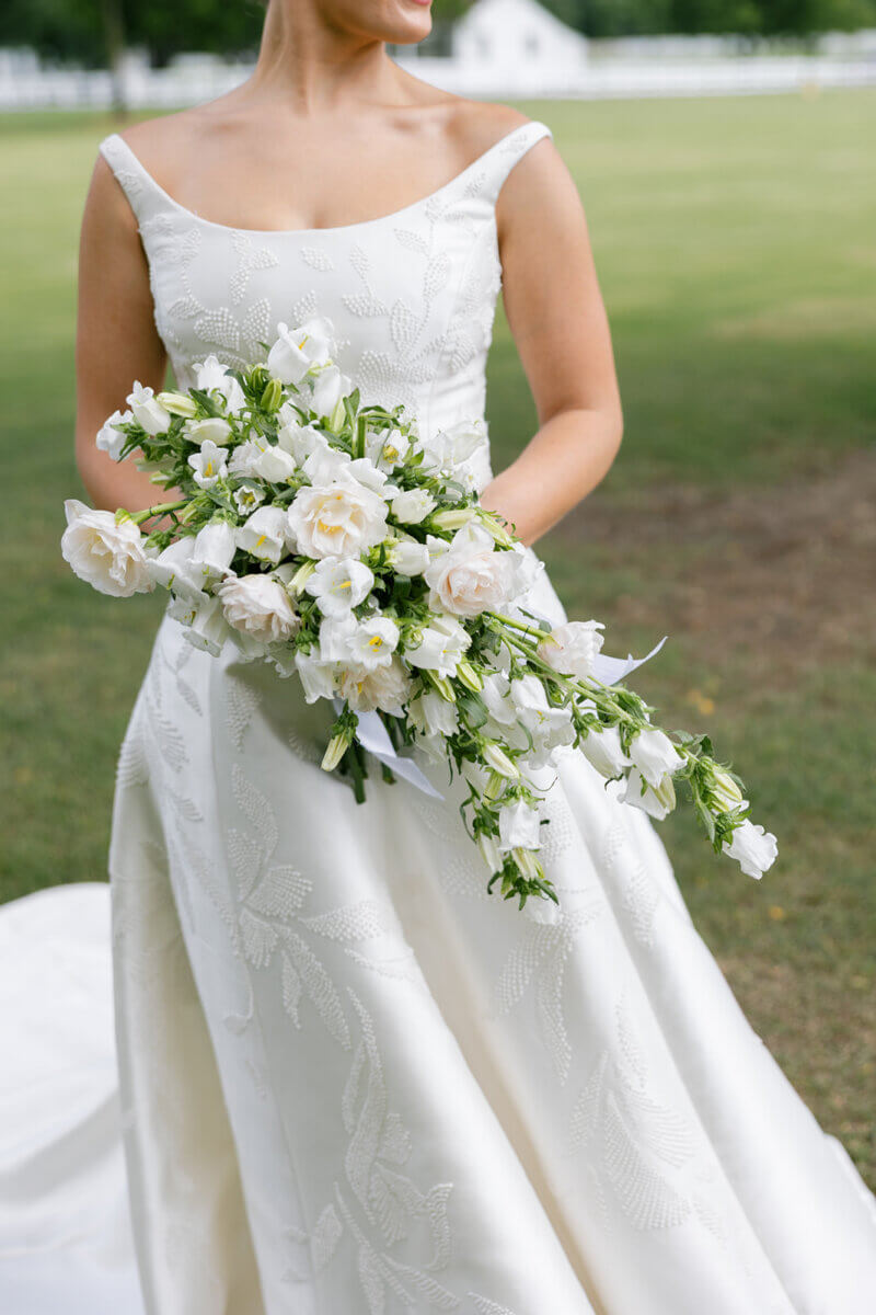 This Dreamy Equestrian Wedding Made History at Franklin's Harlinsdale Farm - 6 A bride in a white embroidered gown holds a cascading bouquet of white and pale pink flowers, standing outdoors on grass.