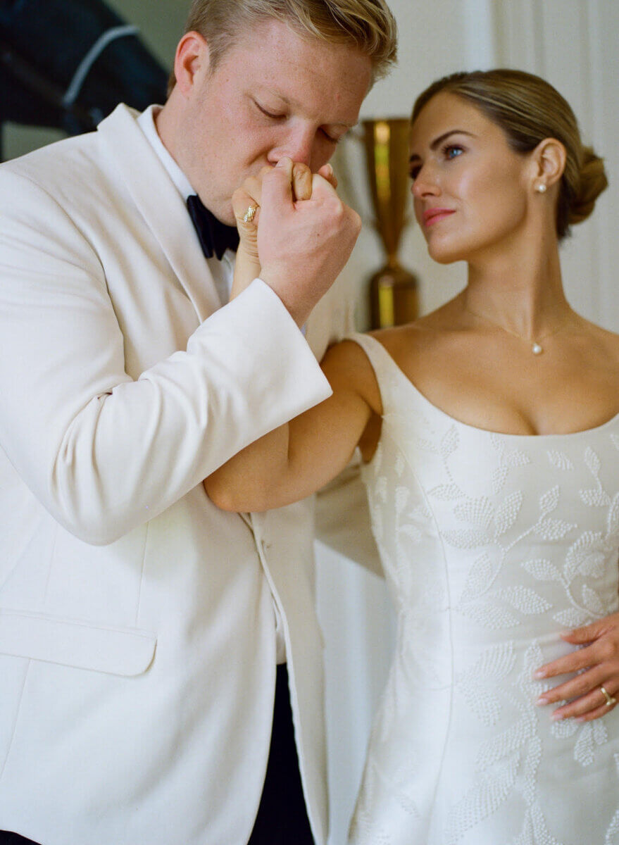 This Dreamy Equestrian Wedding Made History at Franklin's Harlinsdale Farm - 3 A man in a white suit kisses a woman's hand at what appears to be a wedding. The woman, wearing a white dress, looks at him with a slight smile, both dressed elegantly for the special occasion.