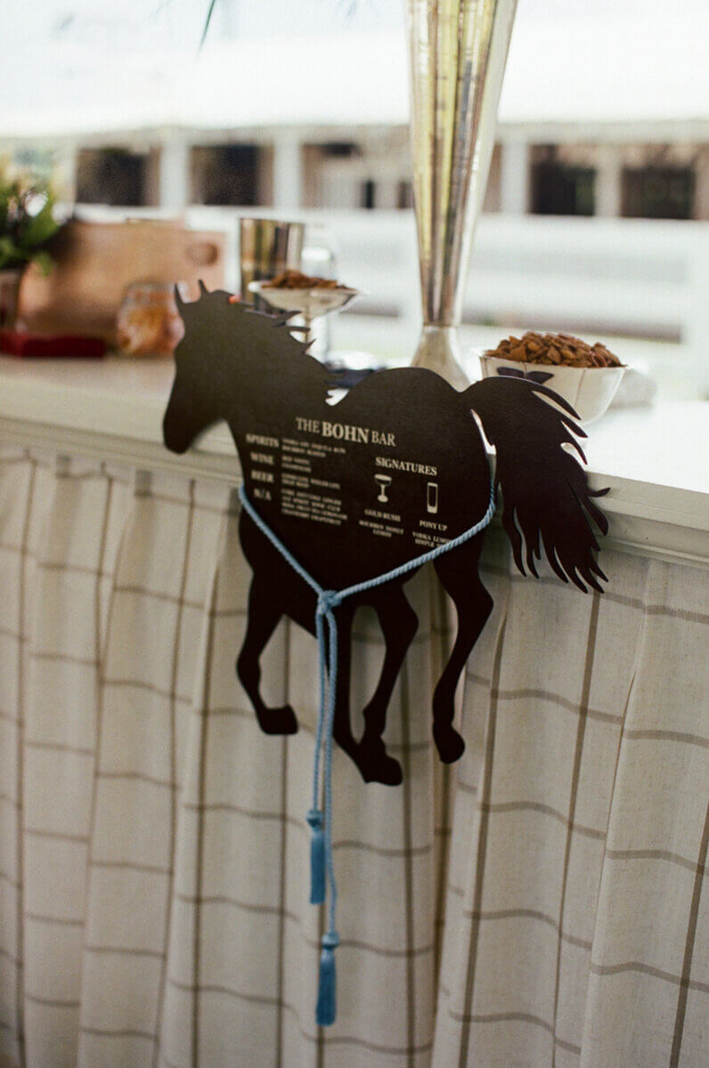This Dreamy Equestrian Wedding Made History at Franklin's Harlinsdale Farm - 11 A black horse-shaped sign displaying a drink menu hangs on a white draped bar at this ethereal wedding venue, with blue cord and bowls of snacks on the counter nearby—a charming detail from a beautiful Franklin wedding.