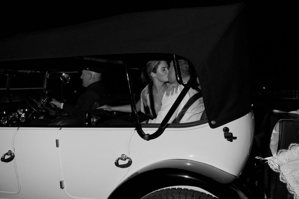 This Dreamy Equestrian Wedding Made History at Franklin's Harlinsdale Farm - 17 A bride and groom sit in the back of a vintage car at night, kissing, while a driver in uniform sits at the wheel, moments after their beautiful Harlinsdale Farm wedding in Franklin.