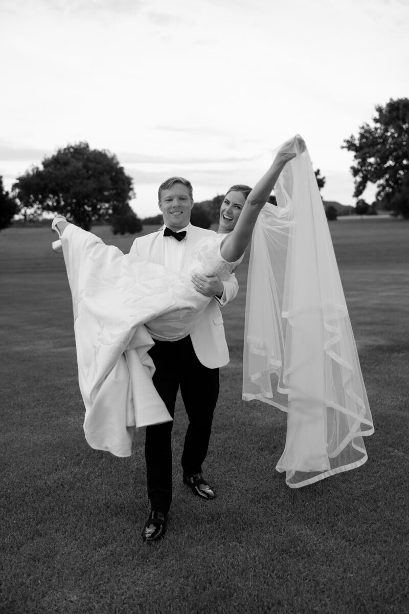 This Dreamy Equestrian Wedding Made History at Franklin's Harlinsdale Farm - 19 A groom in a tuxedo carries a smiling bride in a wedding dress and veil across a grassy field at Harlinsdale Farm, with trees in the background, capturing the joyful spirit of their Franklin wedding.