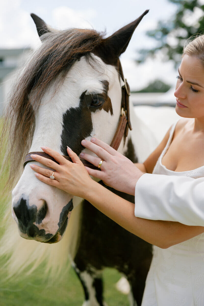 This Dreamy Equestrian Wedding Made History at Franklin's Harlinsdale Farm - 8 A woman in a white outfit gently holds the face of a black and white horse outdoors on a sunny day at Harlinsdale Farm in Franklin, making for a serene wedding-inspired moment.