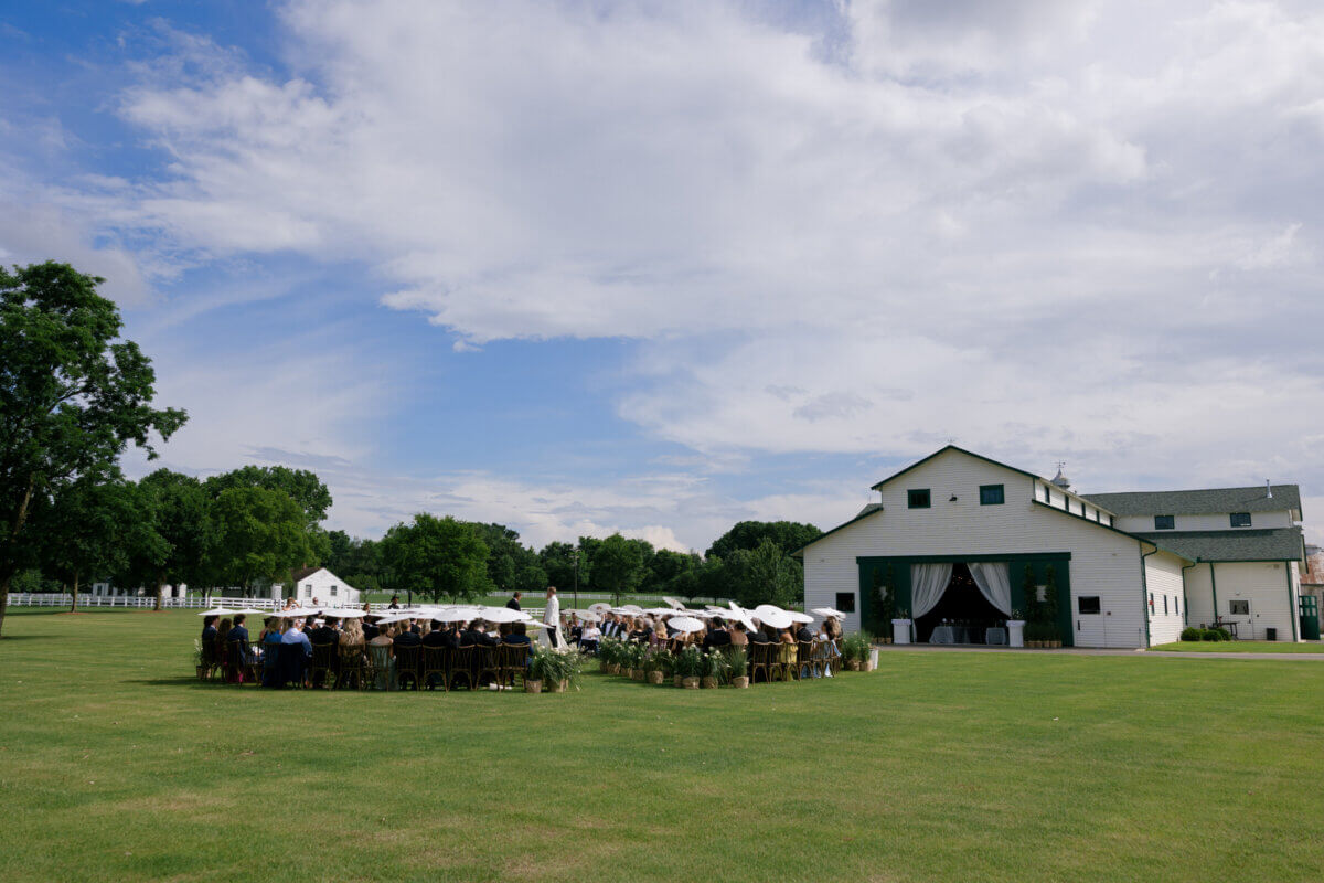 This Dreamy Equestrian Wedding Made History at Franklin's Harlinsdale Farm - 5 A group of people sits in a circle outdoors on a lawn near a large white barn under a partly cloudy sky at Harlinsdale Farm in Franklin, creating an inviting setting perfect for a wedding or special gathering.