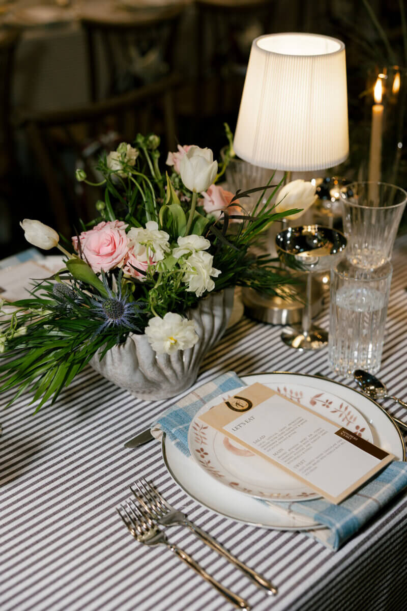 This Dreamy Equestrian Wedding Made History at Franklin's Harlinsdale Farm - 15 Table setting with striped tablecloth, floral centerpiece, lamp, menu on a plate with a blue napkin, fork and knife, and glassware.