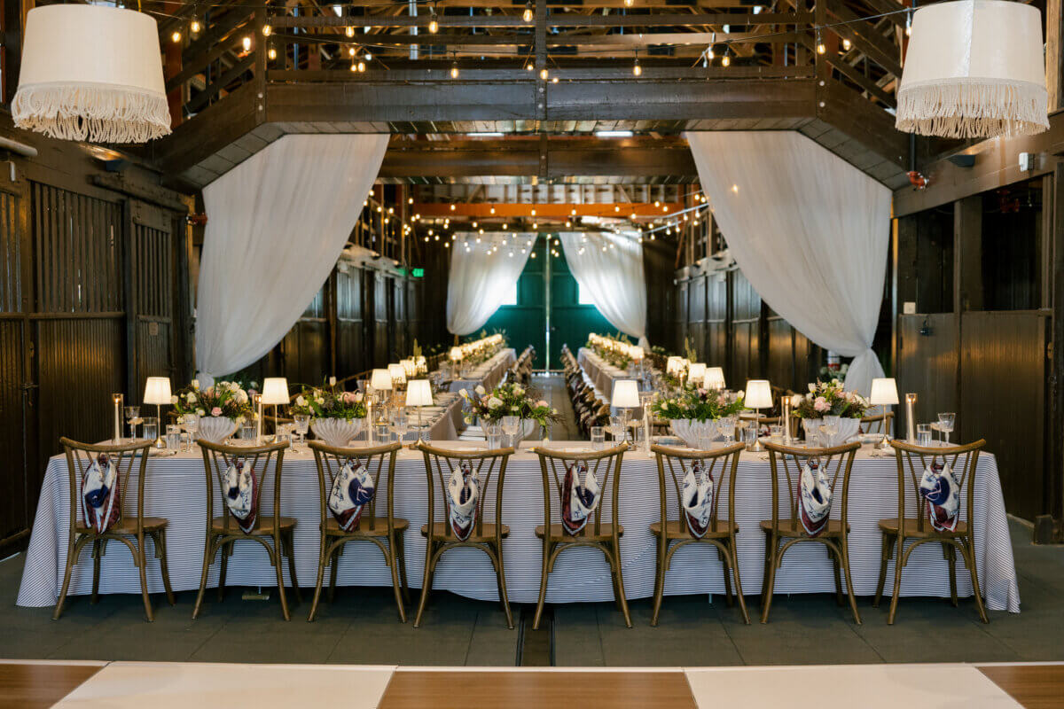 This Dreamy Equestrian Wedding Made History at Franklin's Harlinsdale Farm - 14 Long banquet tables with white tablecloths and decorated chairs are set for a wedding event in a barn at Harlinsdale Farm, Franklin, with string lights and draped white fabric creating an elegant atmosphere.