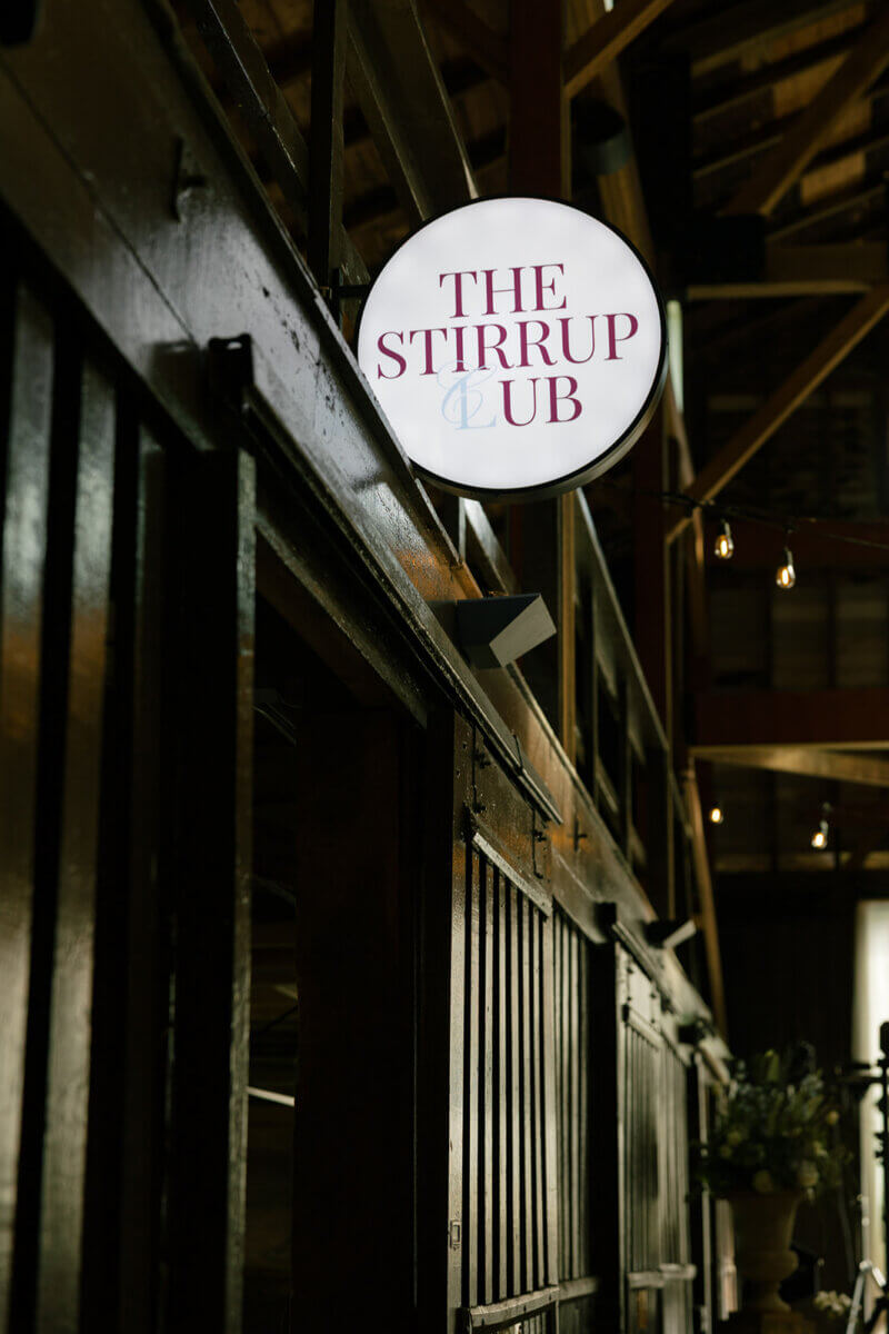This Dreamy Equestrian Wedding Made History at Franklin's Harlinsdale Farm - 16 A round sign reading "The Stirrup Club" is mounted above dark wooden stalls inside a building with exposed beams.