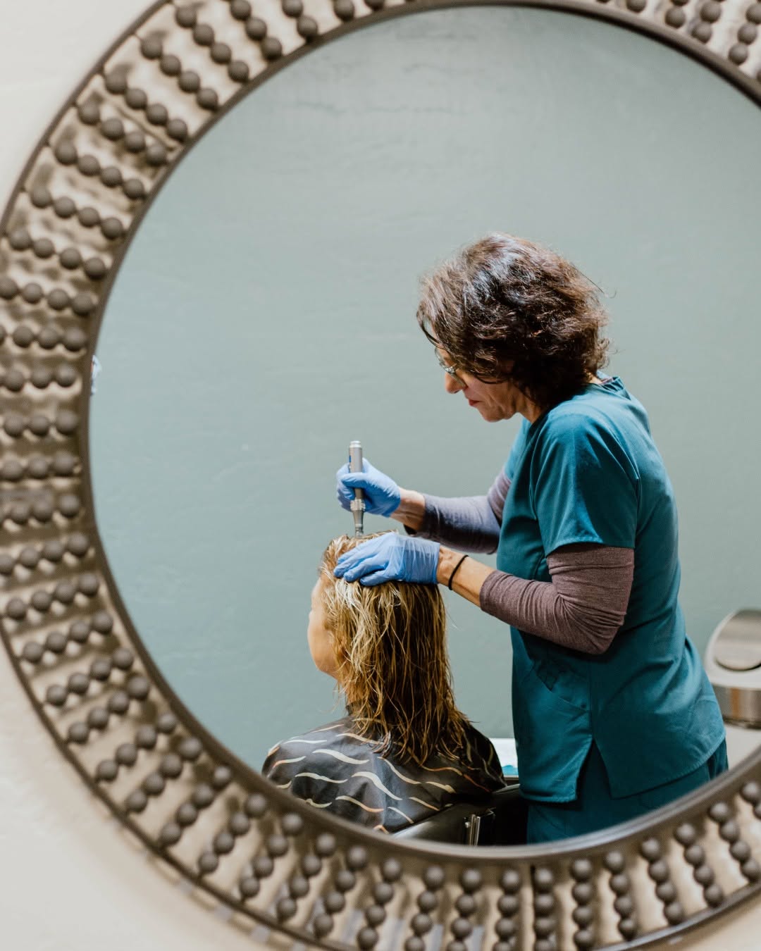 A hairdresser from HPIHair Partners wearing gloves works on a seated client's wet hair, reflected in a round, textured mirror—showcasing the expertise of a leading hair clinic dedicated to hair restoration.