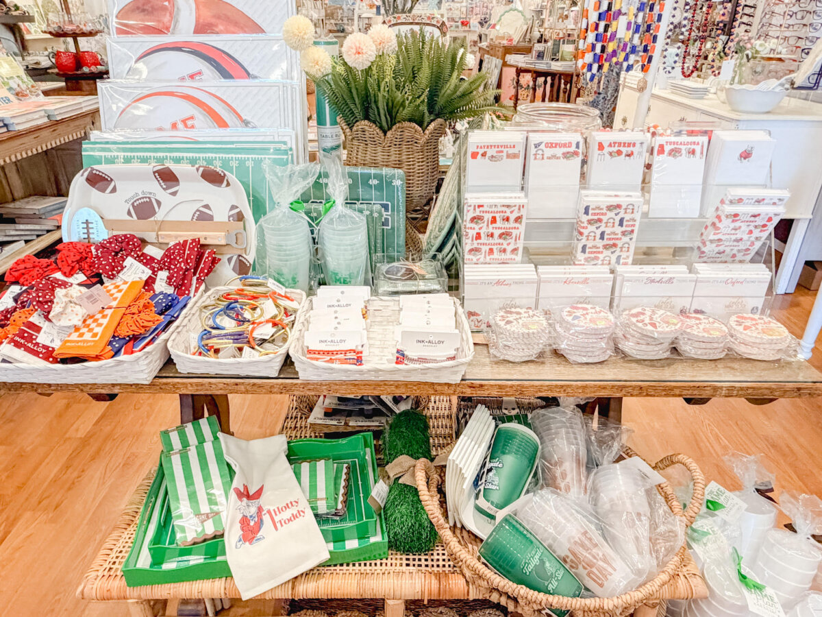 A display table in a gift shop with plates, napkins, drinkware, napkin rings, stationery, and decorative items arranged in baskets and trays on wooden shelves.