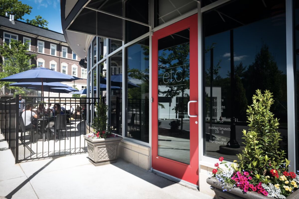 A restaurant entrance with a red door, large windows, and flower planters. Outdoor seating with blue umbrellas and people dining is visible beside the building, making it a perfect spot to catch up on the latest Nashville announcements or discover exclusive Nashville deals.