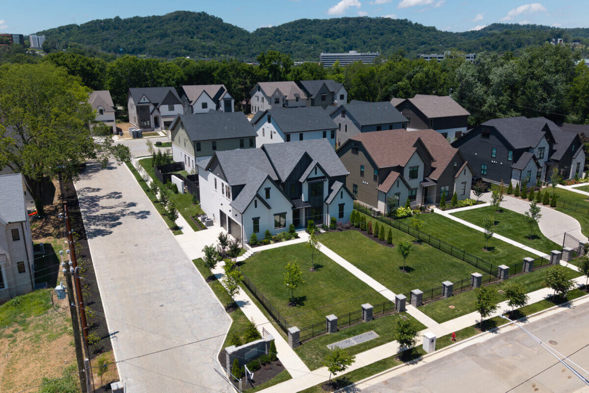 A Look Inside Brentwood's Newest Luxury Address - 2 Aerial view of the newest suburban neighborhood in Brentwood, featuring modern luxury houses, green lawns, sidewalks, and a nearby road, all surrounded by trees and hills in the background.