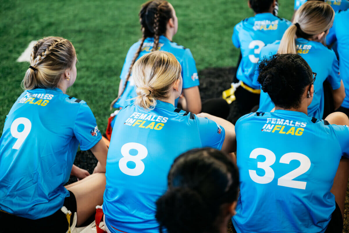 A group of female athletes in blue jerseys with numbers are seated on artificial turf, participating in a Youth Programs flag football event.