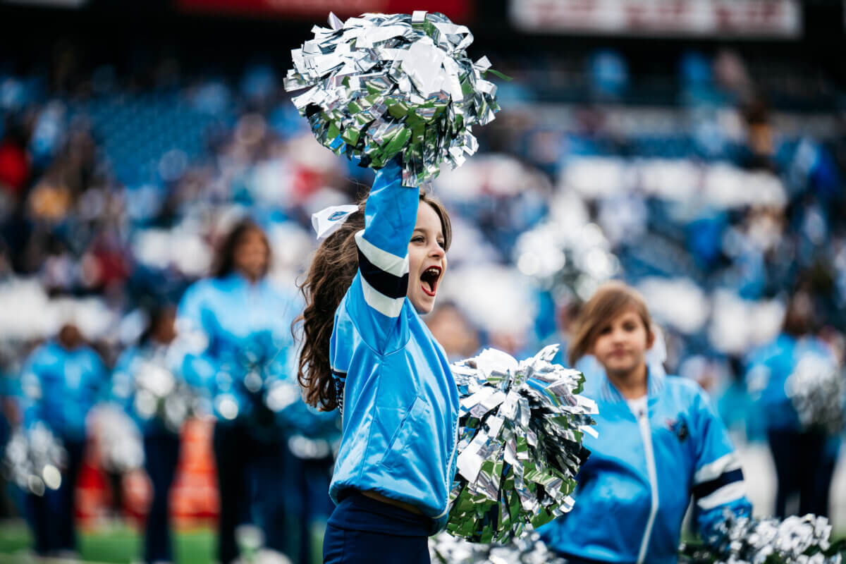 A young cheerleader in a blue uniform holds silver pom-poms and cheers at a stadium, participating in one of the exciting Youth Programs hosted by the Tennessee Titans, with other cheerleaders visible in the background.