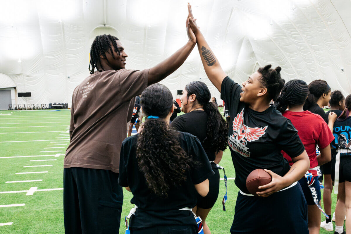 Two people give each other a high five on an indoor football field, surrounded by teammates, with one holding a football—capturing the energetic spirit of Tennessee Titans Youth Programs designed for kids and teens.