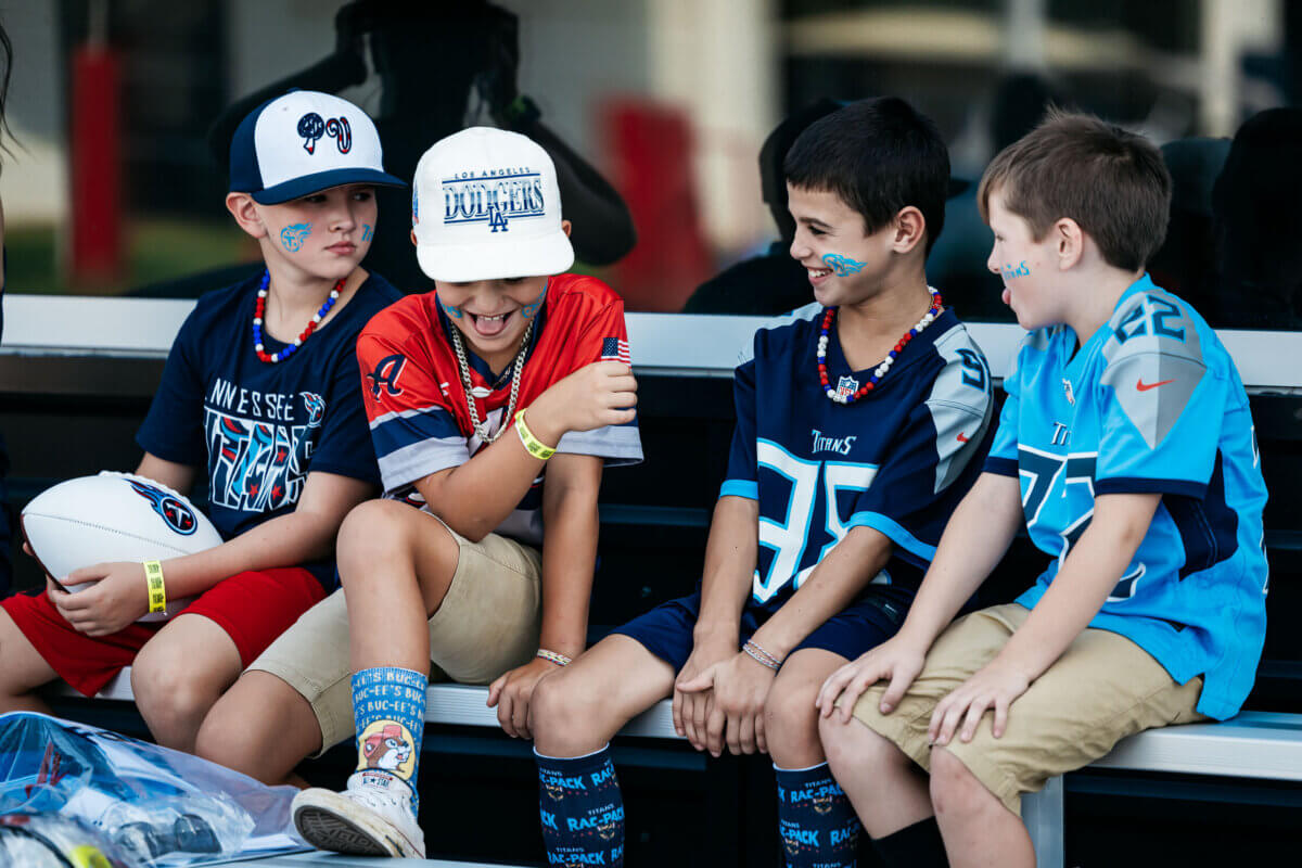 Four boys wearing sports jerseys and hats sit on a bench, smiling and talking, with football-themed face paint and accessories—enjoying one of the fun Youth Programs inspired by Tennessee Titans Kids Experiences.