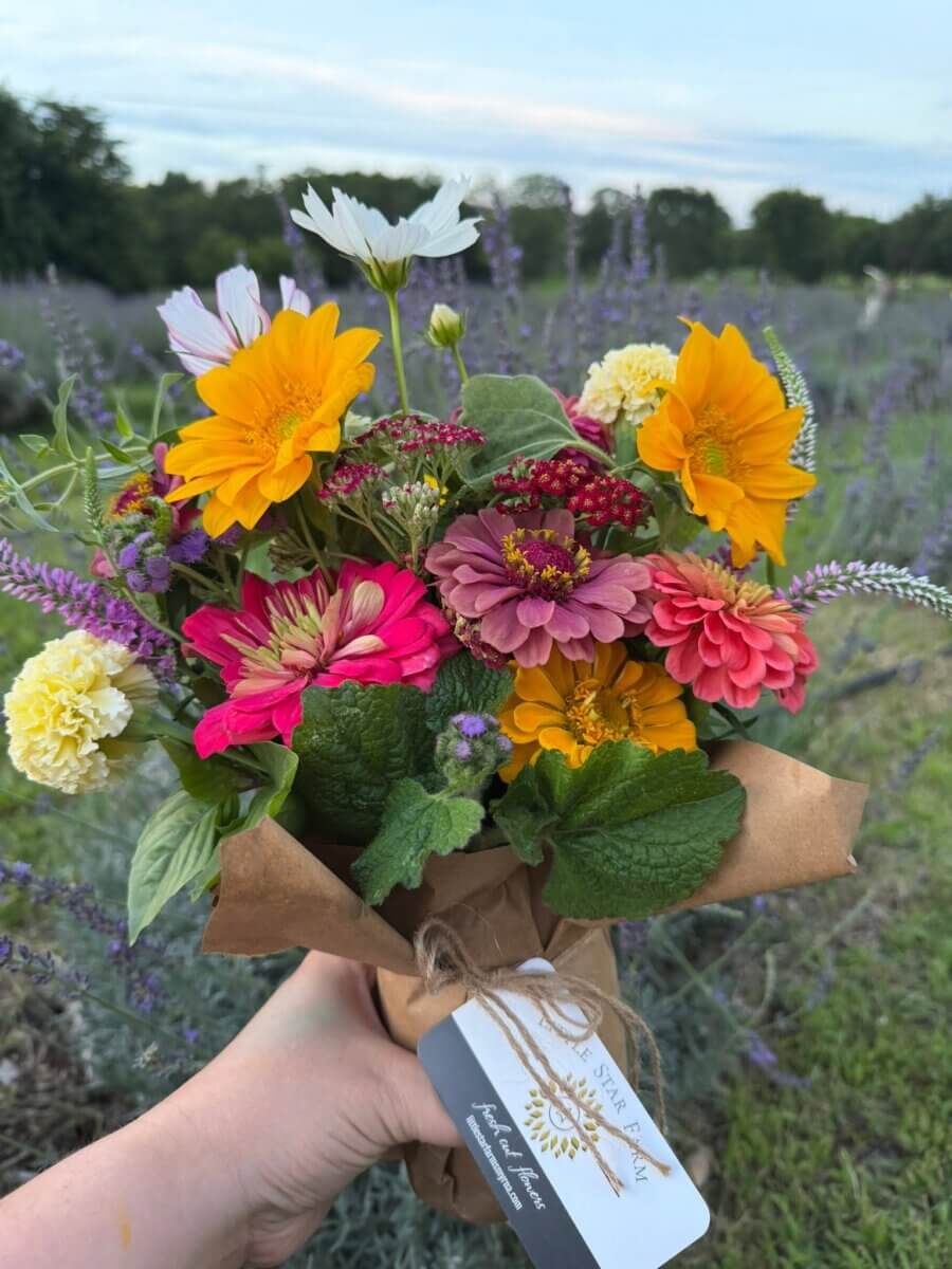 12 Beautiful U-Pick Flower Farms Near Nashville - 5 A hand holds a bouquet of colorful flowers wrapped in brown paper in an outdoor field at a U-Pick Flower Farm near the Nashville area, with a tag attached to the bouquet.