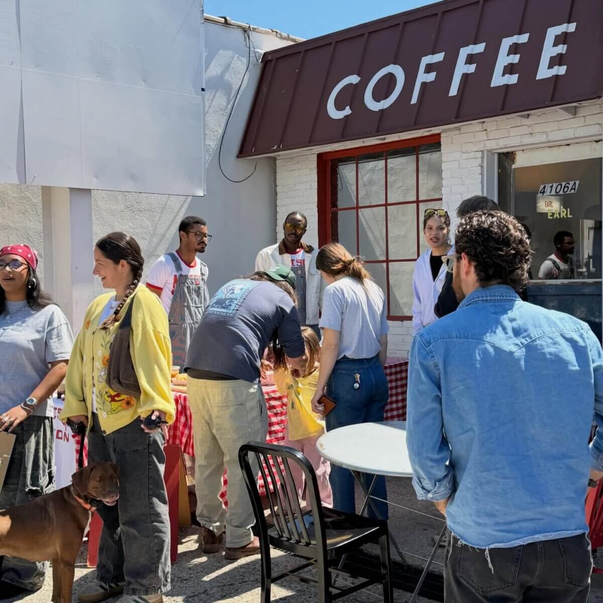 11 Walk-Up Windows To Try in Nashville - 2 A group of people stand and talk outside a coffee shop with a red-and-white checkered table; one person holds a dog on a leash, enjoying the casual atmosphere typical of walk-up restaurants Nashville is known for.