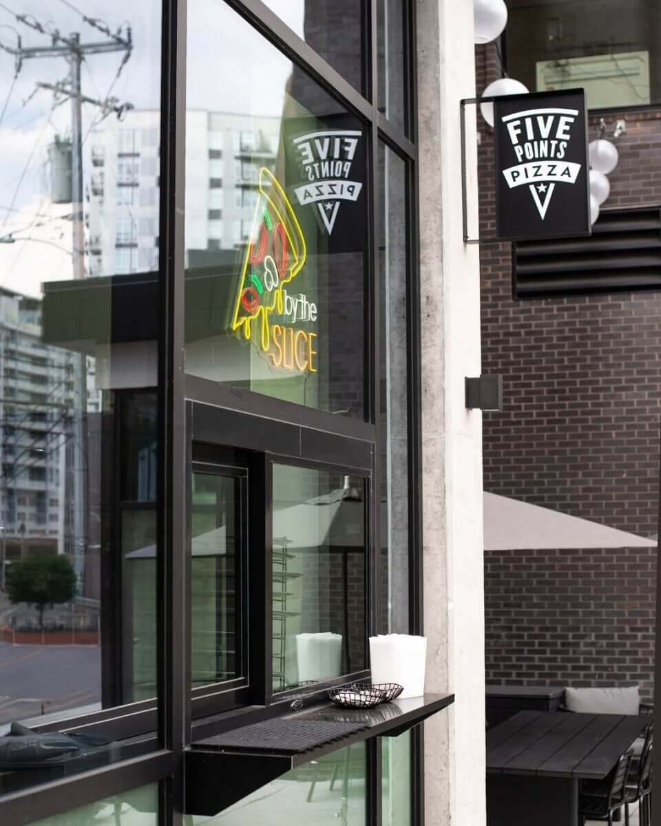 11 Walk-Up Windows To Try in Nashville - 4 Exterior of Five Points Pizza in Nashville with a neon "by the slice" sign, a convenient walk-up window for easy ordering, napkins, and a tip jar; modern buildings and patio seating are visible in the background.