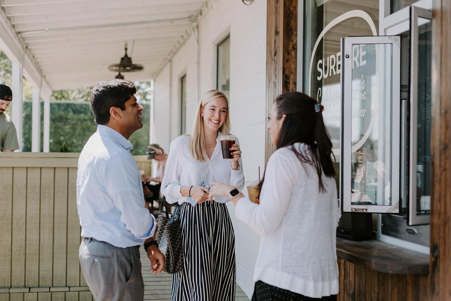 Three people stand outside a building in Nashville, talking and smiling. One woman holds a drink, while the others face her by the walk-up windows. The scene is casual and takes place on a sunlit porch near a window.