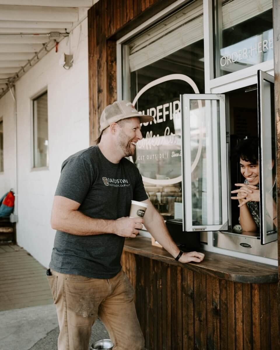 11 Walk-Up Windows To Try in Nashville - 7 A man holding a coffee cup smiles and talks to a woman inside a coffee shop through a drive-thru window, capturing the friendly vibe found at many walk-up windows Nashville is known for.
