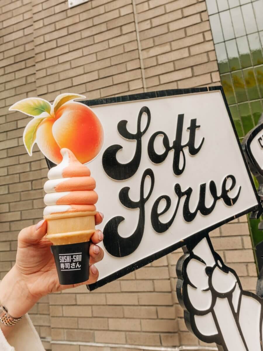 11 Walk-Up Windows To Try in Nashville - 8 Hand holding a peach and vanilla swirl soft serve ice cream cone in front of a "Soft Serve" sign with a peach illustration and brick wall backgroundβone of the sweetest walk-up windows Nashville has to offer.