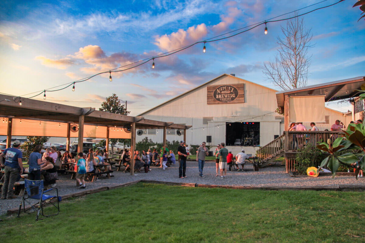 People gather outside a brewery in Natchez MS at sunset, sitting at picnic tables and standing under string lights. A stage and a grassy area are visible in front of the building, adding to the inviting atmosphere for anyone enjoying a weekend in Natchez.