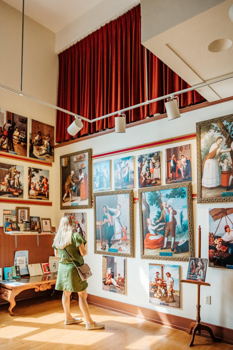 A woman in a green dress stands in an art gallery in Natchez, Mississippi, viewing several framed paintings on a well-lit wall beneath red curtains—a perfect stop during a weekend trip.