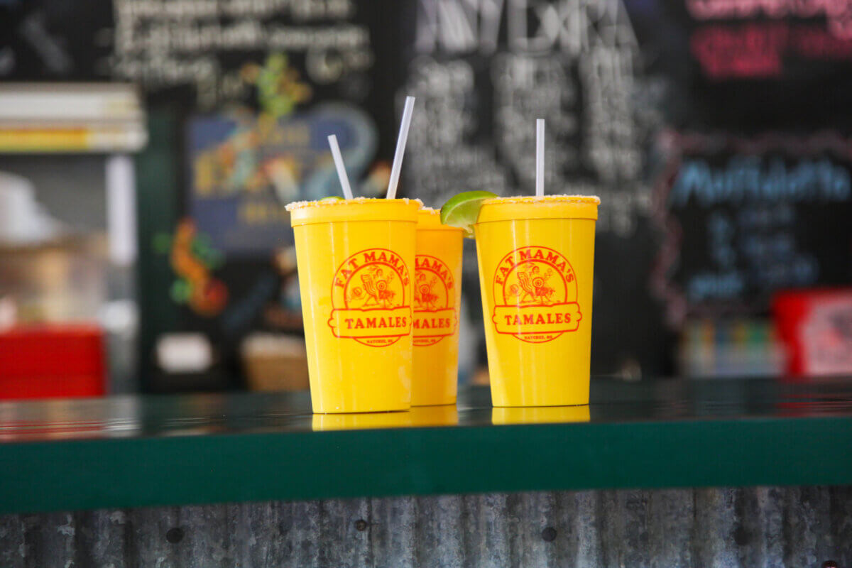 Two yellow plastic cups with "TAMALLES" printed on them are filled with drinks—one garnished with a lime wedge, both with straws—sitting on a counter in Natchez, MS, perfect for a relaxing weekend. The background is softly blurred, letting the vibrant scene take center stage.