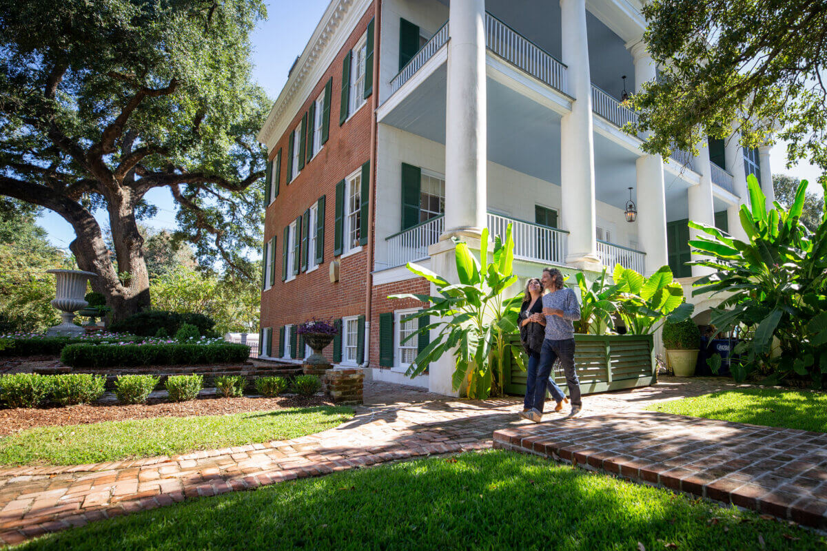 Two people walk on a brick path outside a large historic house with white columns in Natchez, MS, surrounded by green plants and trees on a sunny weekend day.