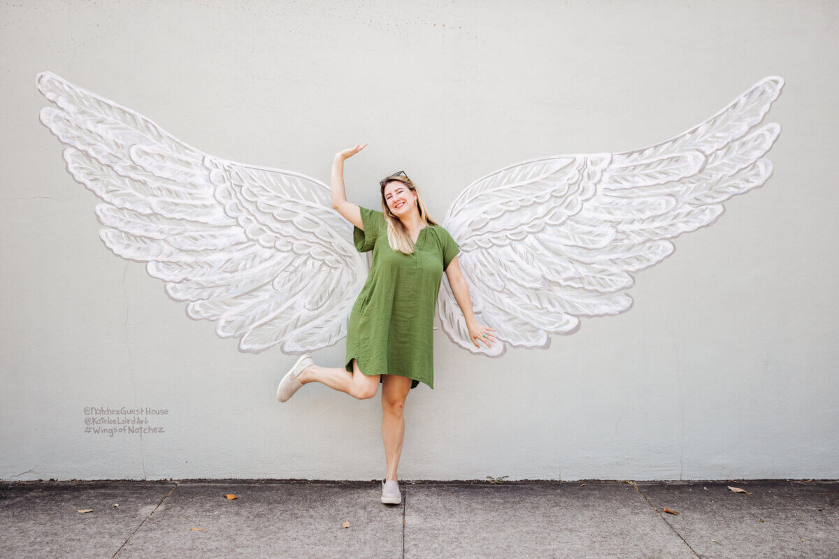 Woman in a green dress poses playfully in front of a mural of large white angel wings on a concrete wall, capturing the perfect travel moment during a weekend adventure in Natchez MS.
