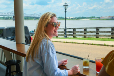 Woman with long blonde hair and sunglasses sits at an outdoor bar overlooking the river with drinks on the counter and a bridge visible in the background.