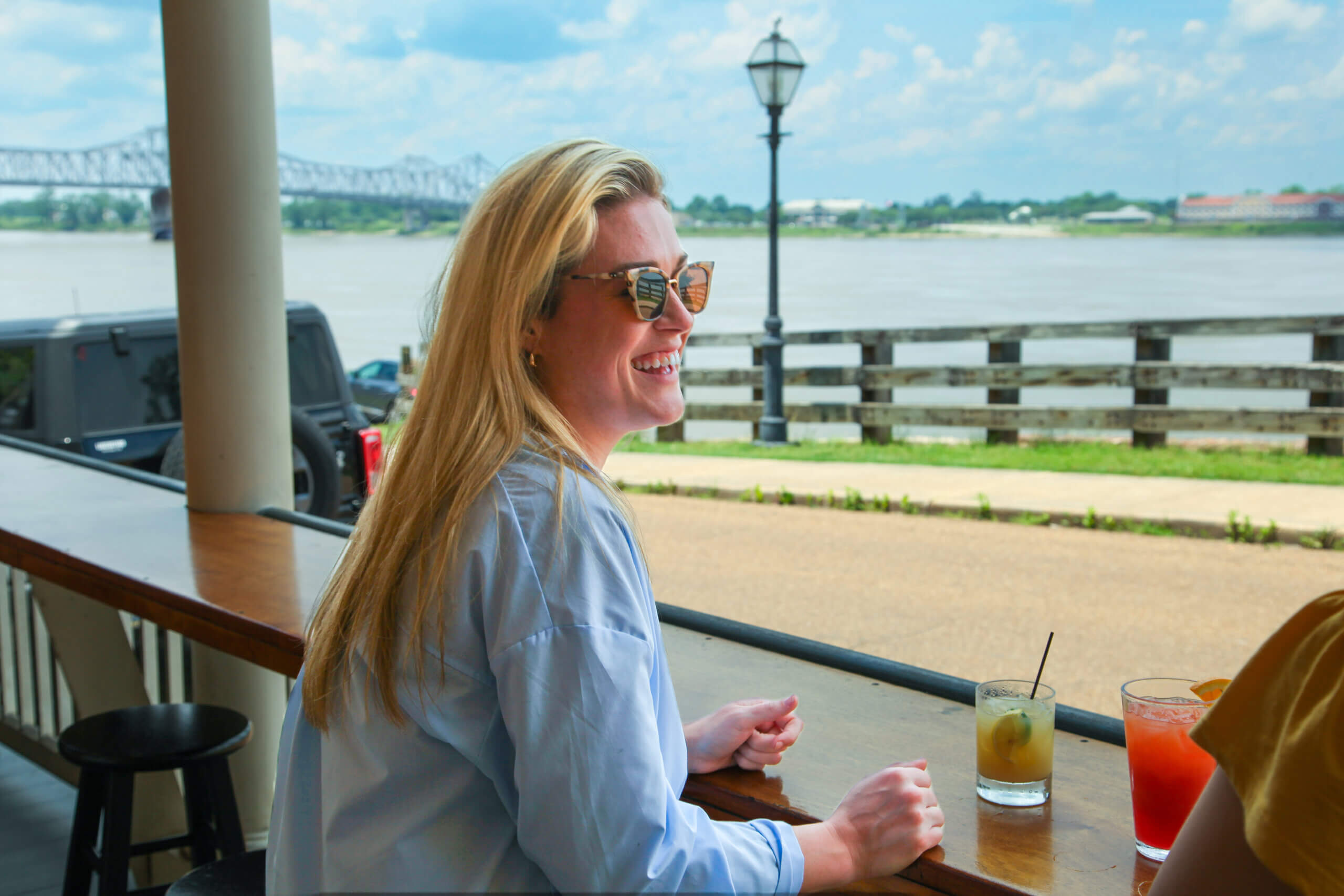 Woman with long blonde hair and sunglasses sits at an outdoor bar overlooking the river with drinks on the counter and a bridge visible in the background.