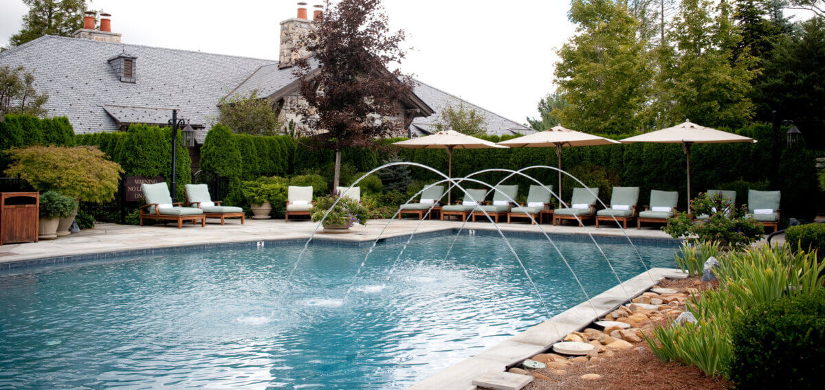 8 Luxurious, Family-Friendly Resorts in the South - 14 Outdoor swimming pool with lounge chairs, umbrellas, and water fountains, surrounded by trees and shrubs, with a large house in the background—perfect for Southern Families seeking relaxing getaways at family-friendly resorts.