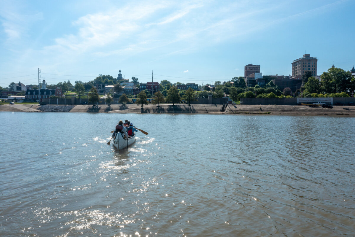 Four people row a small boat on a wide river with a cityscape and trees visible in the background under a clear sky, capturing the spirit of outdoor adventure vacations.