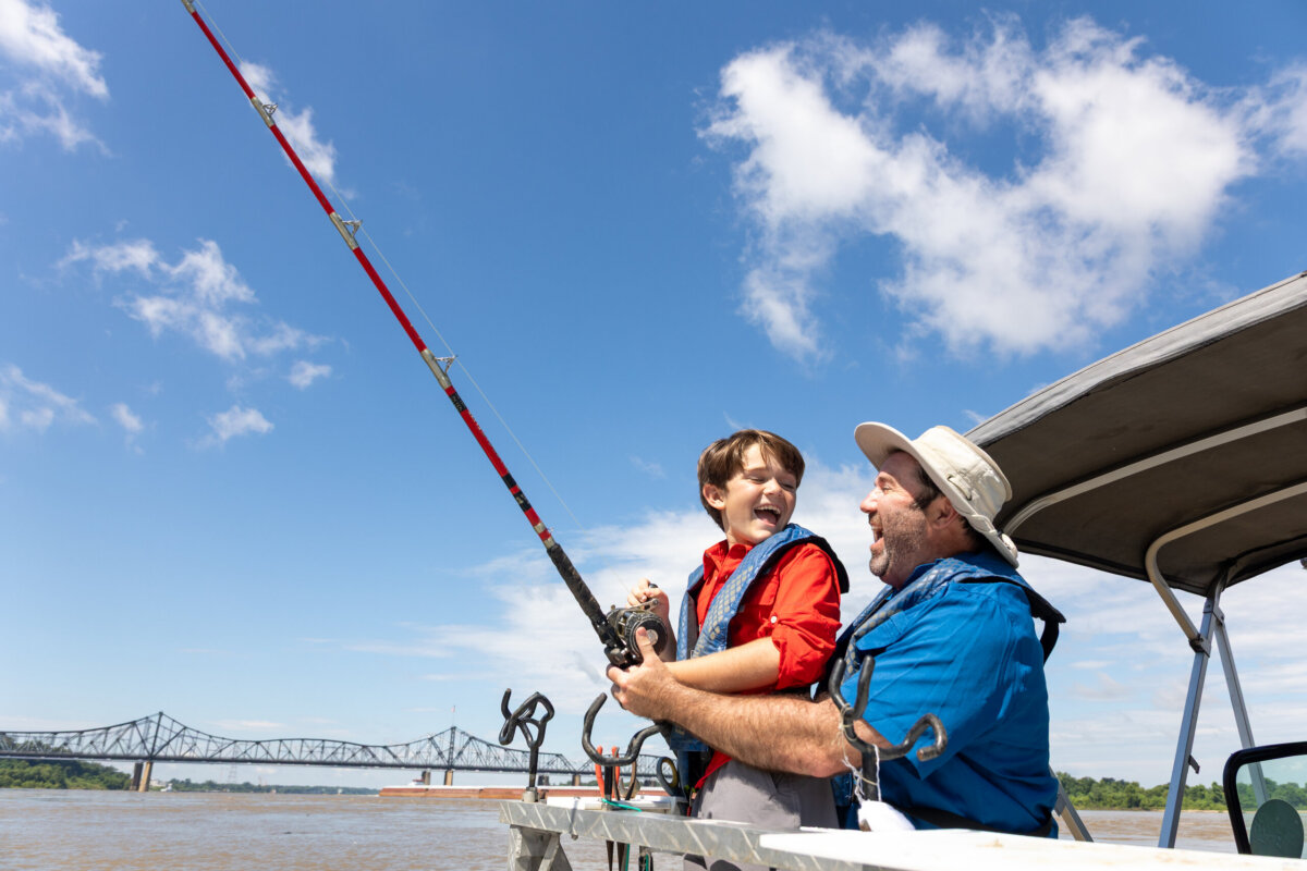 An adult and a child wearing life jackets fish together on a boat under a blue sky, with a bridge visible in the background—capturing the spirit of outdoor adventure vacations.