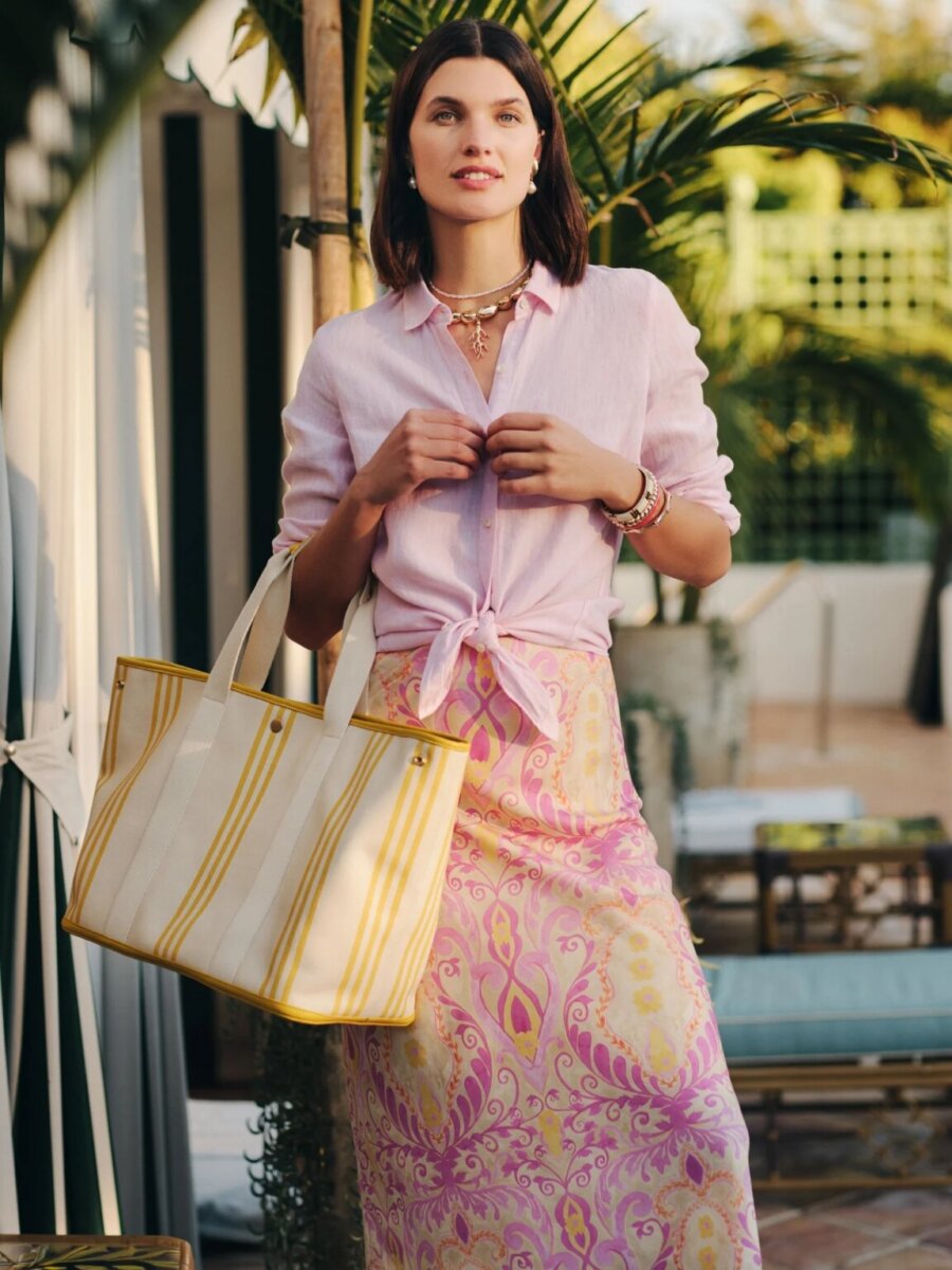 Woman stands outdoors showcasing timeless style in a light pink button-down shirt paired with a pink patterned skirt, holding a large striped tote bag. There are palm trees and patio furniture in the background, highlighting one of the effortless ways to wear button-down outfits during warm-weather days.