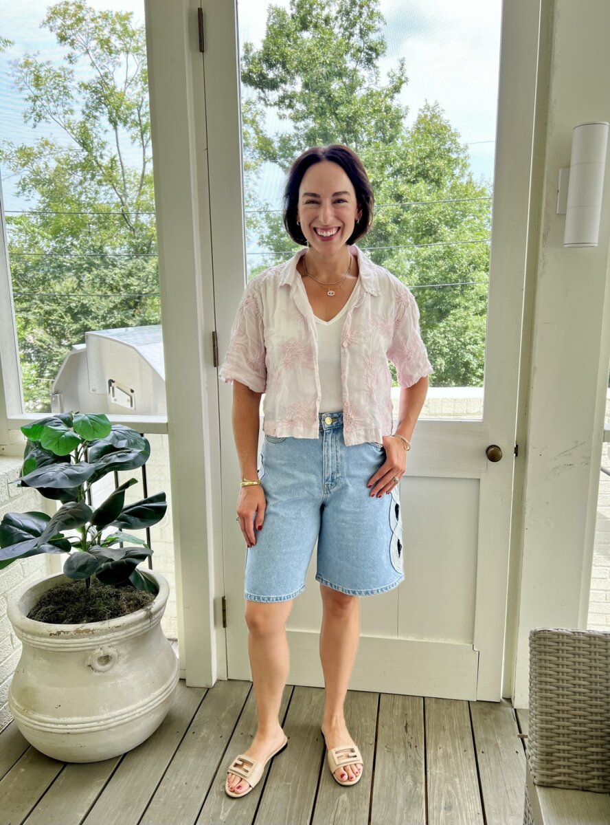 Woman stands on a porch showcasing effortless style, wearing a white top, light pink button-down shirt, denim shorts, and sandals. She is smiling, with one hand in her pocket. A potted plant is visible to her left.