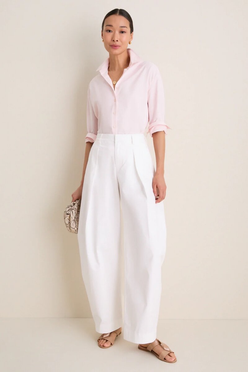 Woman stands against a light wall showcasing her effortless style in a pale pink button-down shirt, white high-waisted wide-leg trousers, tan sandals, and holding a small textured clutch.