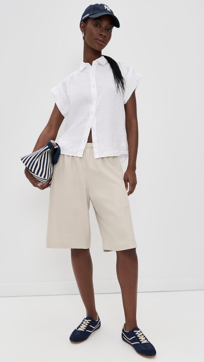 Person showcasing effortless fashion style in a white short-sleeve button-down shirt, beige knee-length shorts, navy sneakers, and a navy baseball cap, holding a navy-and-white striped clutch against a plain white background.