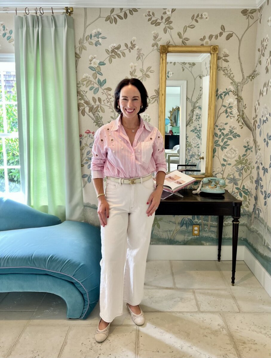 A woman stands smiling in a decorated room with floral wallpaper, a blue chaise lounge, a black table with books, and a gold-framed mirror—showcasing one of the season’s chic button-down outfit ideas.