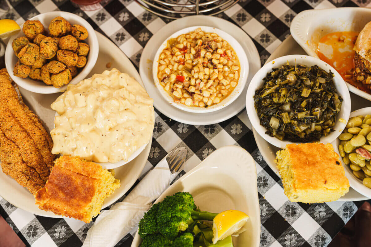 An Ode to Oxford: How to Eat Your Way Through This Delicious Small Town - 20 A table at one of the renowned Oxford restaurants is adorned with dishes including fried okra, creamed corn, greens, lima beans, broccoli, cornbread, catfish fillet, and a plate of potato salad—all beautifully arranged on a black and white checkered tablecloth.