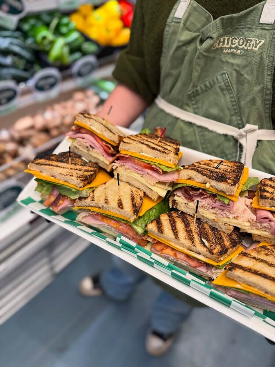 An Ode to Oxford: How to Eat Your Way Through This Delicious Small Town - 19 A person in a green apron holds a tray of grilled sandwiches stacked with cheese, lettuce, and deli meats in a grocery store or market setting.