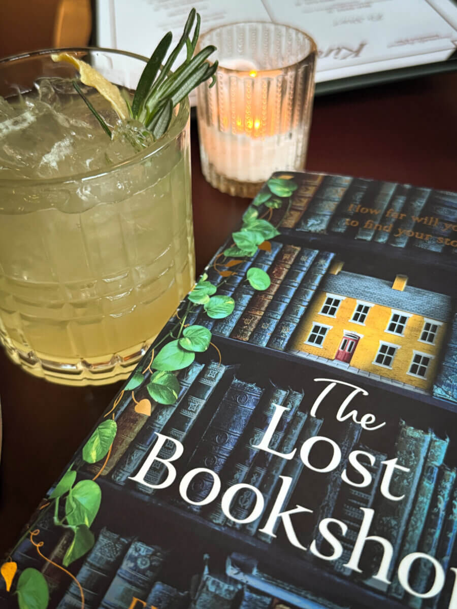 An Ode to Oxford: How to Eat Your Way Through This Delicious Small Town - 3 A glass of cocktail with rosemary garnish sits beside a book titled "The Lost Bookshop" on a table with a lit candle in the background, evoking the cozy ambiance often found in charming Oxford restaurants.
