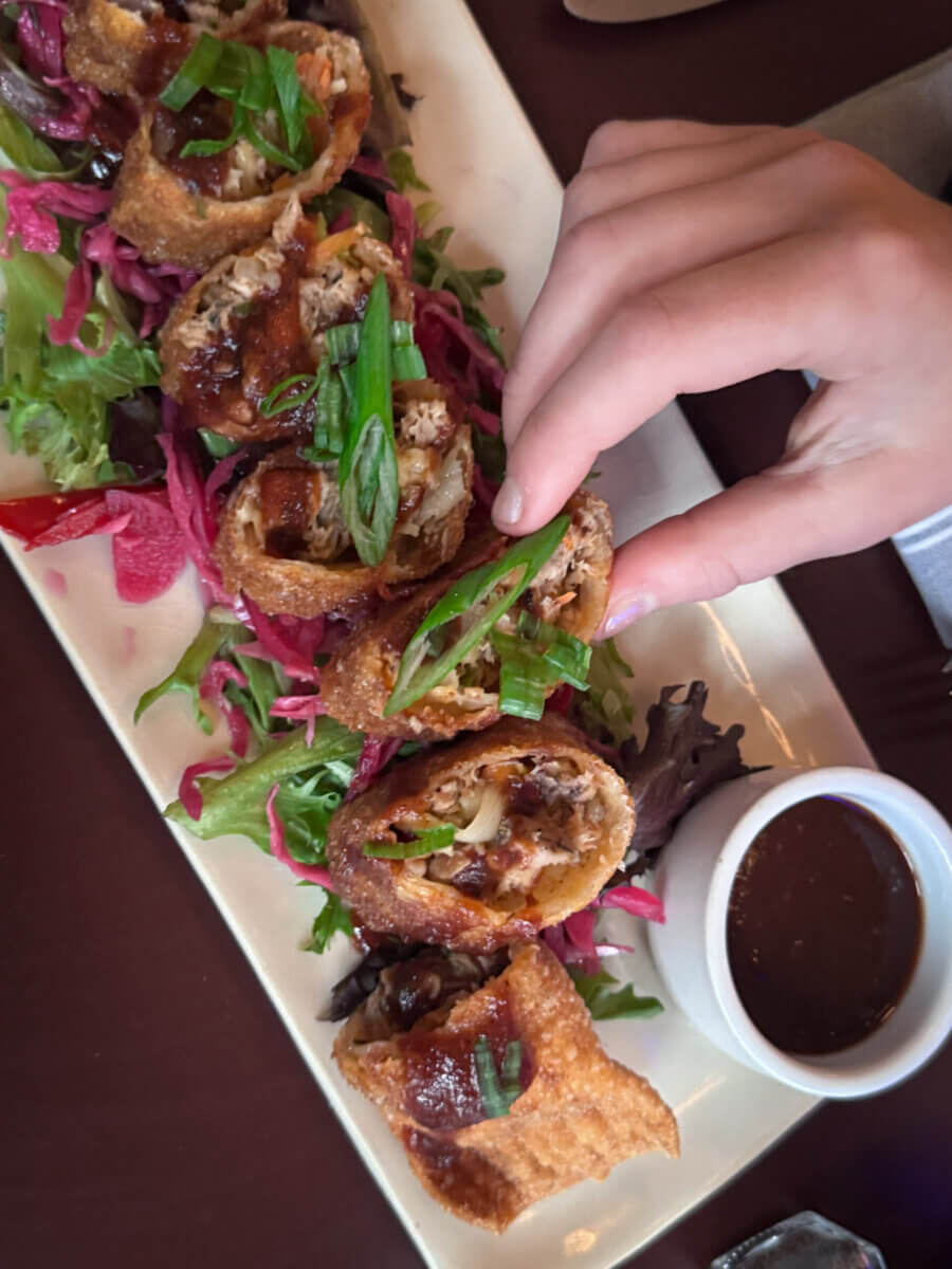 An Ode to Oxford: How to Eat Your Way Through This Delicious Small Town - 4 A hand reaches for a crispy rolled appetizer sliced on a bed of mixed greens, with a small bowl of dipping sauce on the side—a tempting dish.