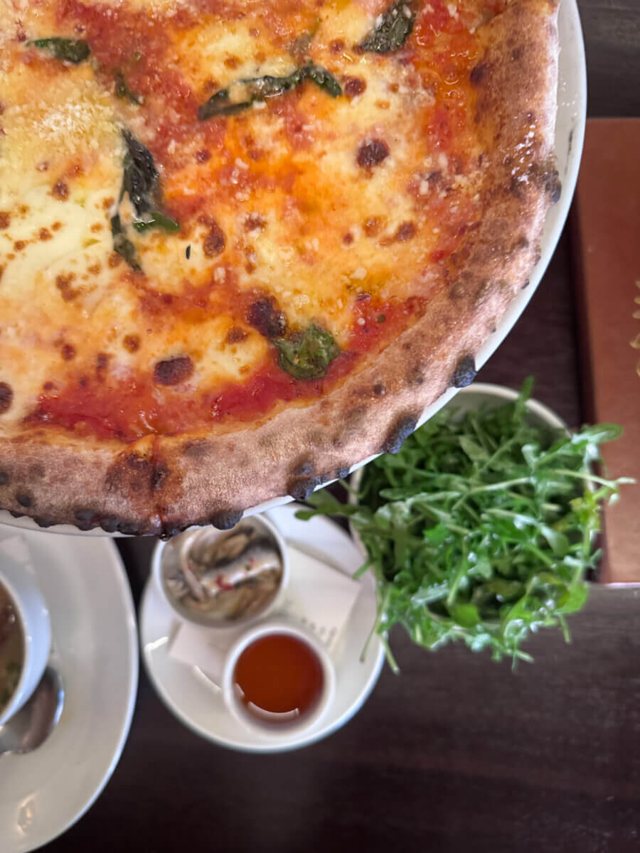An Ode to Oxford: How to Eat Your Way Through This Delicious Small Town - 17 A close-up of a margherita pizza on a plate, with arugula, a small dish of oil, and a side plate with condiments visible on a dark wooden table.