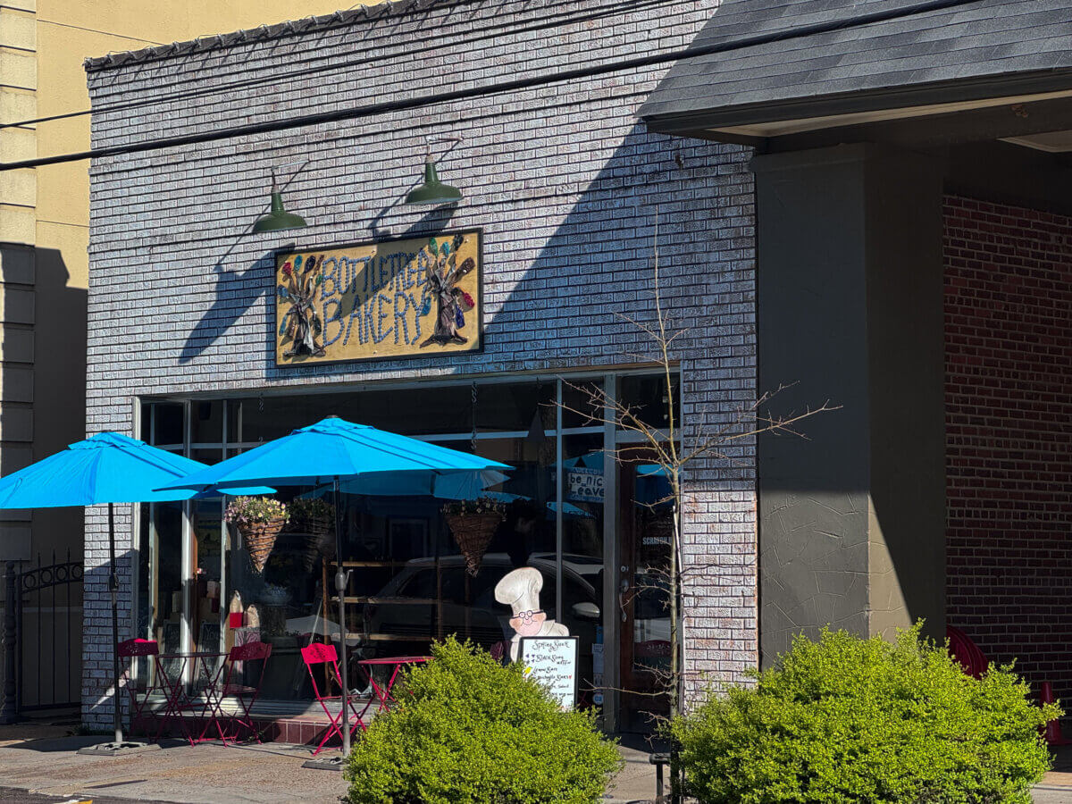 An Ode to Oxford: How to Eat Your Way Through This Delicious Small Town - 15 A small bakery with a brick exterior, blue umbrellas over outdoor tables, a hanging sign reading "bakery," and green bushes in front, reminiscent of the charming style found at many Oxford restaurants.