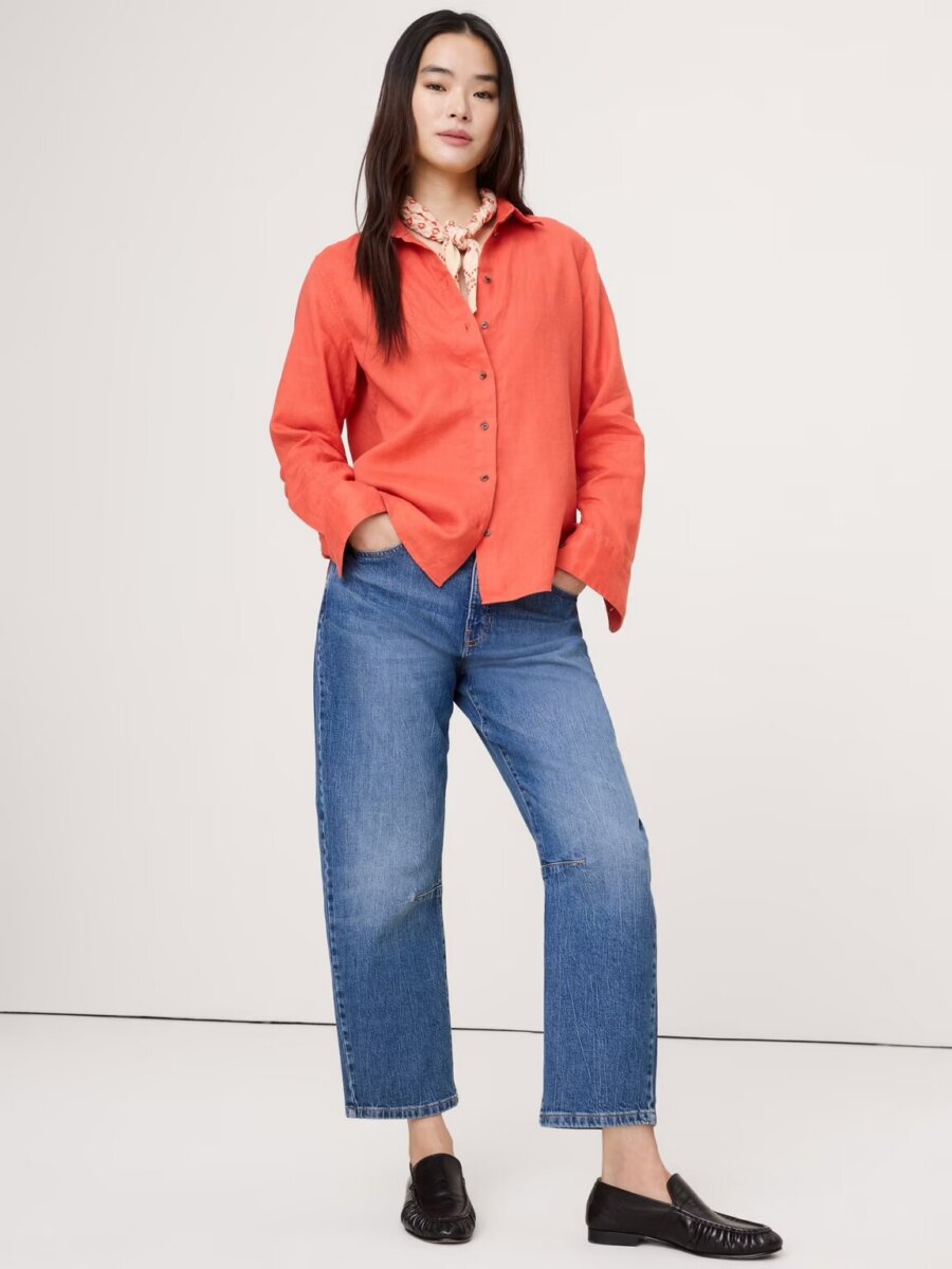 A woman stands against a plain background wearing a coral button-up shirt, blue jeans, a patterned scarf, and black loafers. She faces forward with one hand in her pocket, effortlessly showcasing one of the classic ways to style button-down outfits for any occasion.