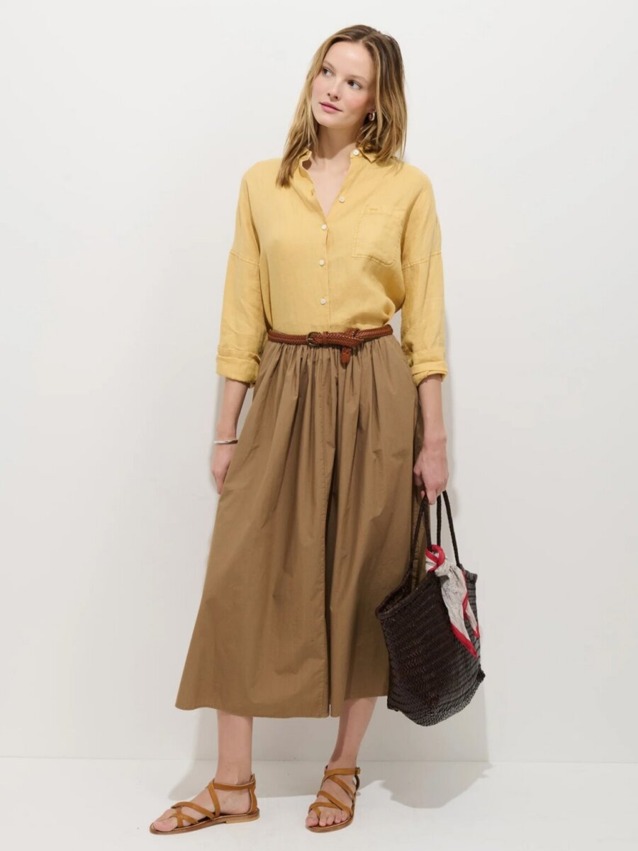 Woman stands against a white background showcasing effortless fashion, wearing a yellow button-down shirt, tan midi skirt, brown belt, and sandals. She holds a woven black tote bag with items inside.